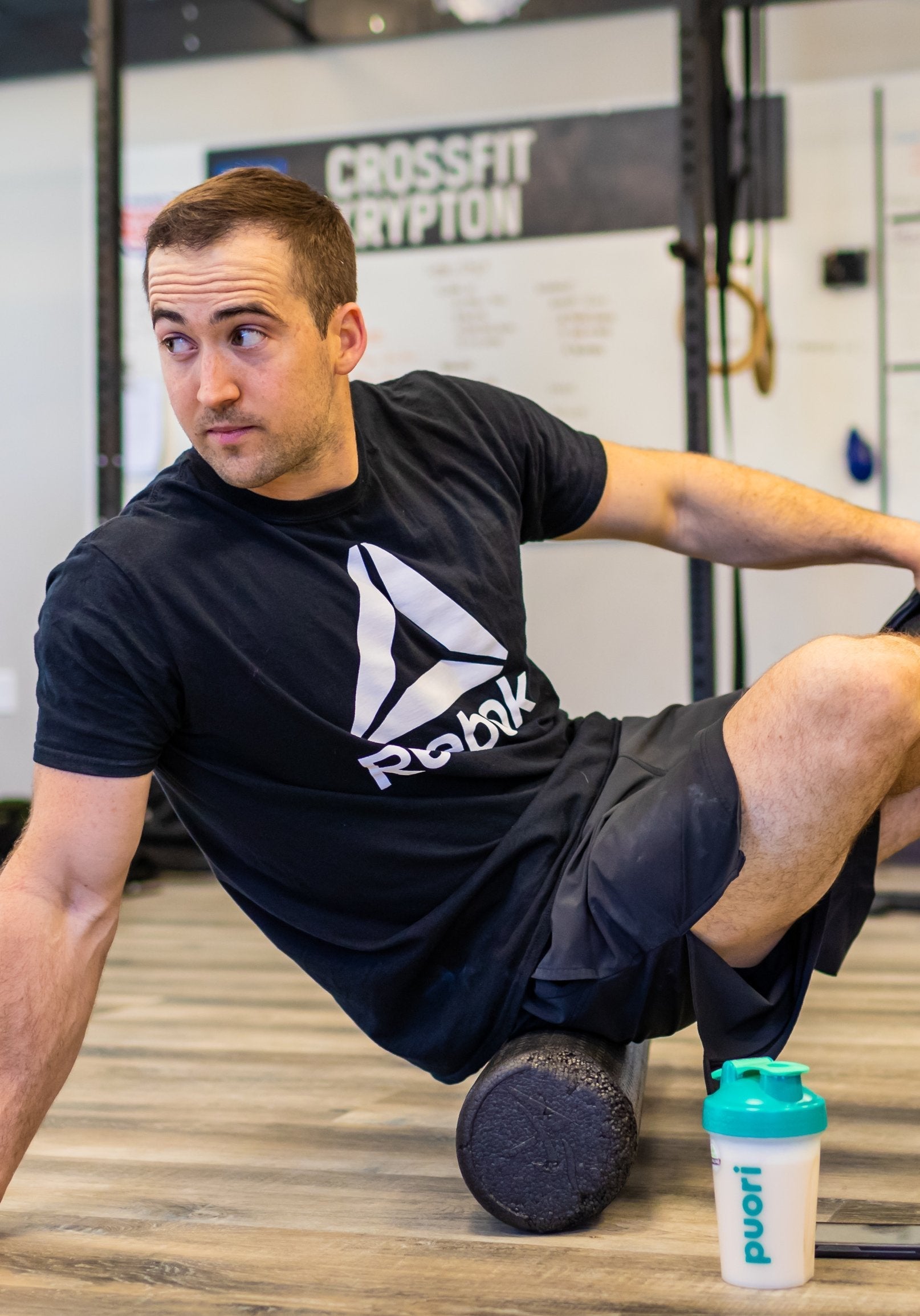 Man in a gym doing a side-plank-style stretch on a foam roller under his leg, with a teal Puori shaker bottle on the floor beside him; CrossFit sign visible in background.